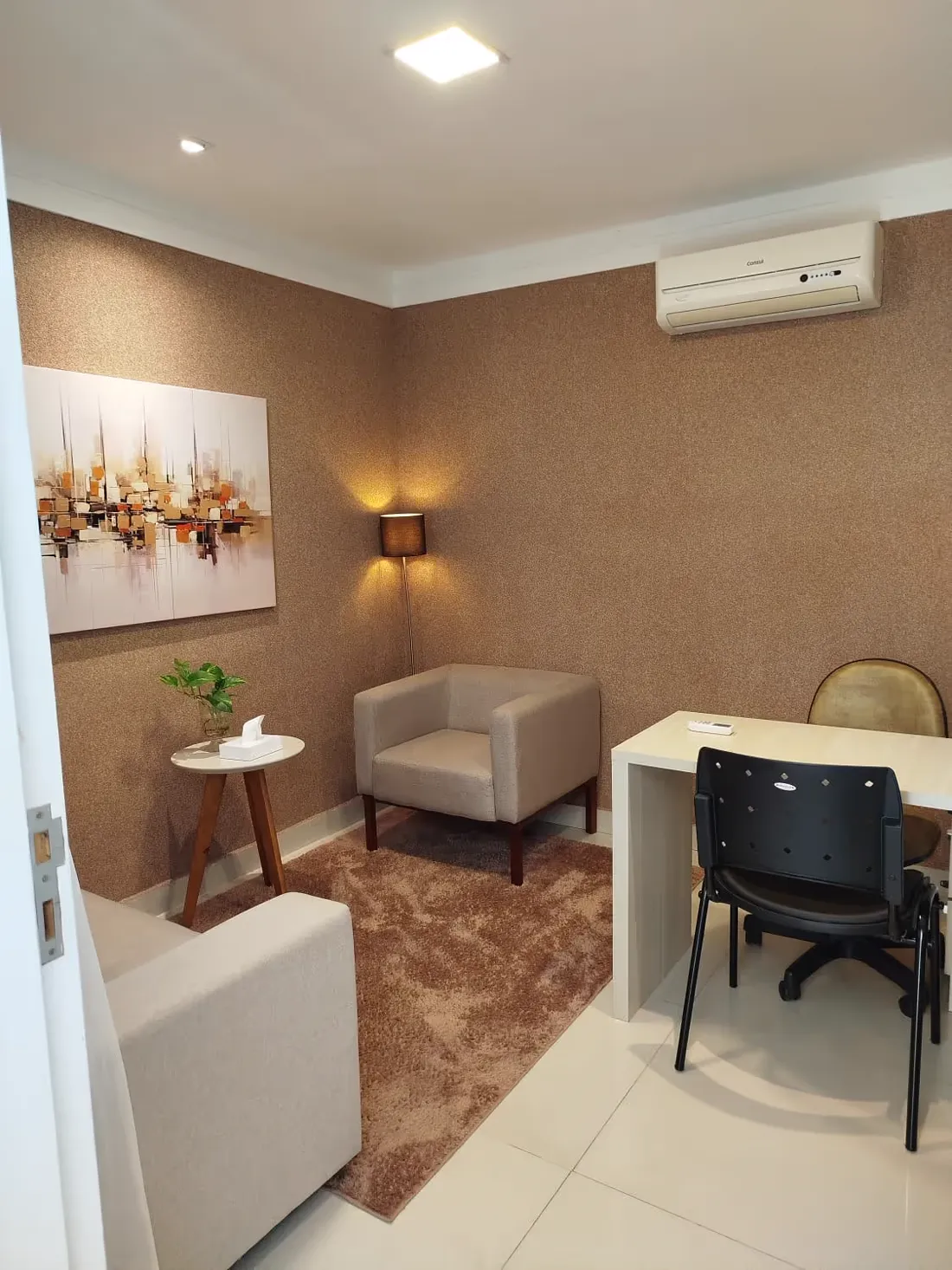 Consultório com poltrona clara, mesa e quadro de tons terrosos